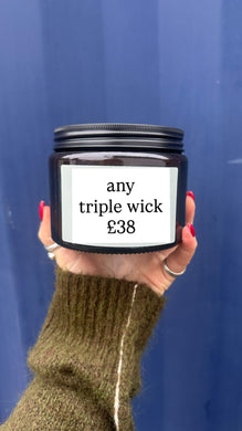 Hand holding a candle jar with a label indicating 'any triple wick £38' against a blue background