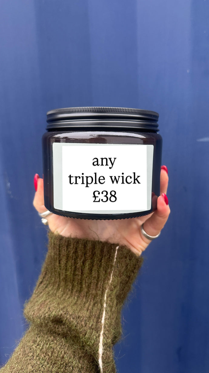 Hand holding a candle jar with a label indicating 'any triple wick £38' against a blue background
